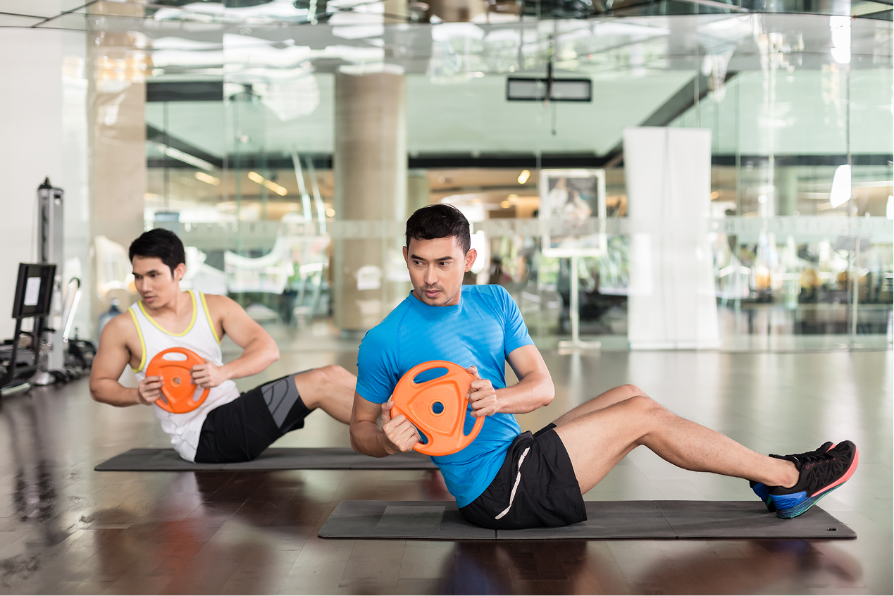Two men doing core exercises with weight plates on mats in gym.